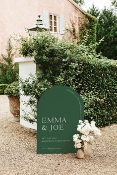 a green sign sitting on top of a gravel ground next to a bush and potted plant