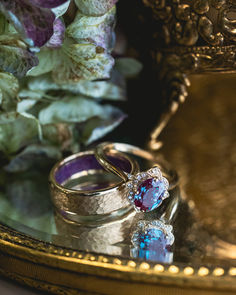 A matching wedding ring set from Staghead Designs sits on a mirror surrounded by flowers and gold trinkets. This ring set features our "Jane" floral engagement ring in 14K yellow gold with an oval lab-grown alexandrite center stone and diamond accents, paired with a 6mm width men's wedding band in hammered 14K yellow gold with sleepy lavender opal lining. Pearl And Alexandrite Ring, Dark Opal Engagement Ring, Vintage Alexandrite Engagement Rings, Purple Rings Engagement, Lavender Wedding Ring, Alexandrite Engagement Rings, Hammered Gold Wedding Band, Opal Wedding Set, Opal Wedding Ring Set