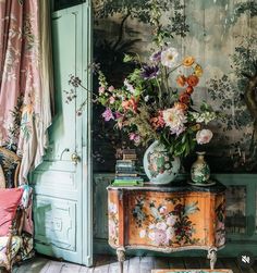 a table with flowers on it in front of a wallpapered background and chair