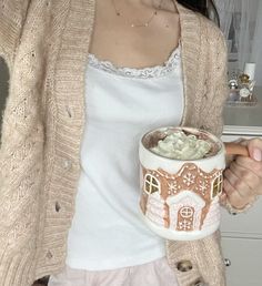 a woman holding a cup with whipped cream in it and a gingerbread house on the mug