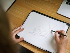 a person is drawing on a paper with a pencil while sitting in front of a laptop