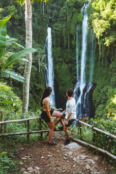 two people are sitting on a railing looking at a waterfall in the jungle, while another person is taking a photo