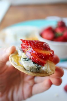 a person holding up a small cracker with fruit on it in front of a bowl of strawberries