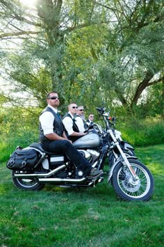 two men are sitting on a motorcycle in the grass with trees behind them and one man is wearing sunglasses