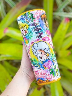 a hand holding up a colorful tube with cartoon characters on it and plants in the background