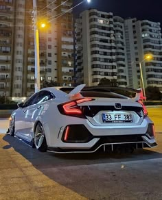 the rear end of a white sports car parked in front of tall buildings at night