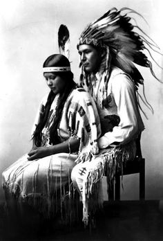 an old black and white photo of two native american indians sitting on a chair with feathers in their hair