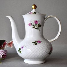 a white tea pot with pink flowers on it next to a flower vase and a rose