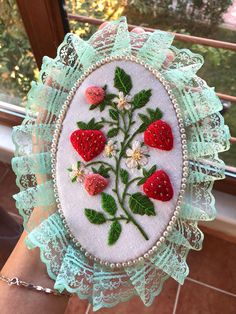 a hand holding up a white and green embroidered badge with strawberries on it, in front of a window