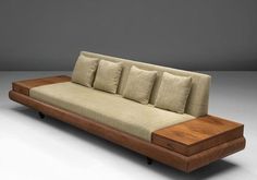 a wooden couch with four pillows on it's back and two sides, sitting in front of a gray wall