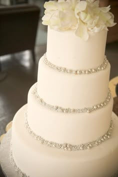 a three tiered white wedding cake with flowers on the top and beaded trim