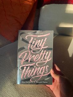 a person holding up a book in their hand on a couch with the title tiny pretty things written on it