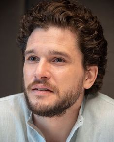 a man with curly hair and beard wearing a blue shirt looks at the camera while he stares into the distance