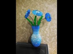 three blue flowers in a vase on top of a black box against a beige wall