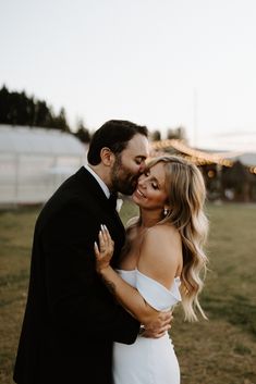 a bride and groom embracing in a field at sunset with lights on the horizon behind them