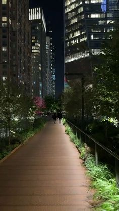 people walking down a sidewalk in the city at night with tall buildings and trees on both sides