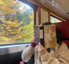 a woman reading a book while sitting on a train in front of a tree filled window