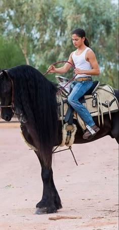 a woman riding on the back of a black horse in a dirt field next to trees