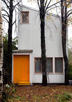 an orange door sits in front of a white building with trees and leaves on the ground