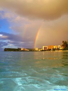 a rainbow is seen over the ocean with buildings in the background and water below it