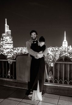 a man and woman standing on top of a roof at night with the city lights in the background