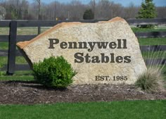 a large stone sign sitting in front of a fenced in area with grass and bushes