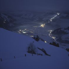 a snowy mountain with lights in the distance and snow on the ground at night time