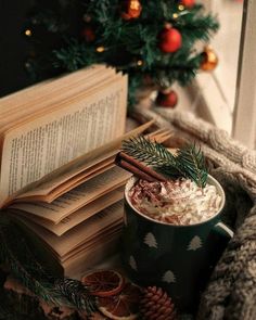 an open book and cup of hot chocolate with cinnamon on the table next to christmas tree