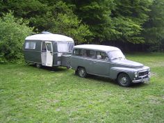 an old gray van and trailer parked in the grass near some trees with green leaves