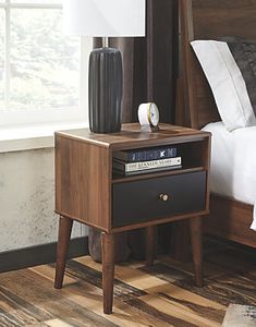 a night stand with a lamp on top of it and a bed in the background