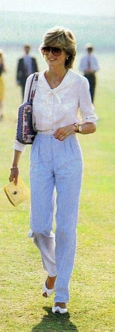 a woman walking across a grass covered field holding a purse and wearing white clothing with people in the background