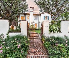 a white house surrounded by trees and flowers on a brick path leading to the front door