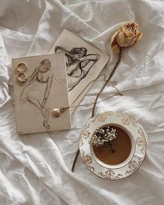 a cup of tea next to two drawings and a rose on a white bed sheet