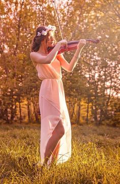 a woman in a pink dress playing the violin on a grassy field with trees behind her