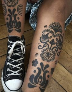 a woman's legs with tattoos on them sitting on a wooden floor next to a pair of sneakers