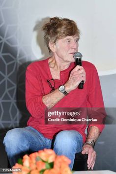 an older woman sitting in front of a microphone and talking into a microphone with flowers on the table behind her