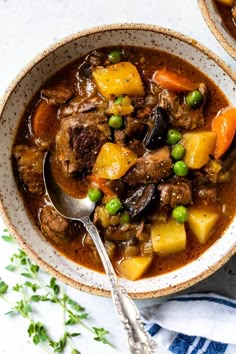 two bowls of beef stew with potatoes and carrots on a white tablecloth next to a spoon