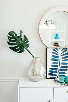 a white dresser topped with a mirror and a vase filled with a green palm leaf