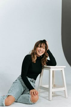 a smiling woman sitting on the ground in front of a white stool with her legs crossed