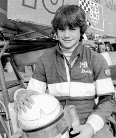 a young man sitting in front of a race car holding a helmet