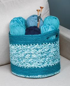 a crocheted basket with yarn and knitting needles in it on a couch next to a pillow