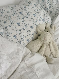 a white teddy bear laying on top of a bed next to pillows and sheets with blue flowers