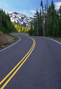 an empty road in the mountains with trees and snow capped mountain in the back ground