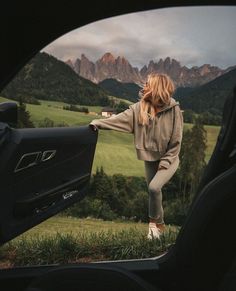 a woman standing next to a car looking out the window at mountains and trees in the distance