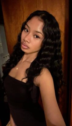 a woman with long black hair standing in front of a wooden wall and looking at the camera