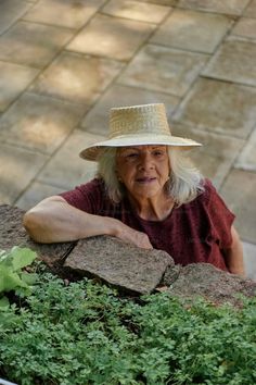 an older woman wearing a straw hat leaning against a rock