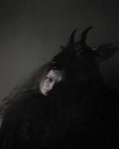 a woman with long hair wearing a horned mask and black coat, in front of a dark background