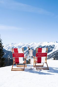 two red and white lawn chairs sitting on top of a snow covered slope with mountains in the background