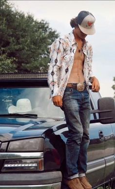 a shirtless man standing on the hood of a truck wearing cowboy boots and a hat