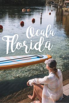 a woman sitting on a dock next to a body of water with a surfboard in the background